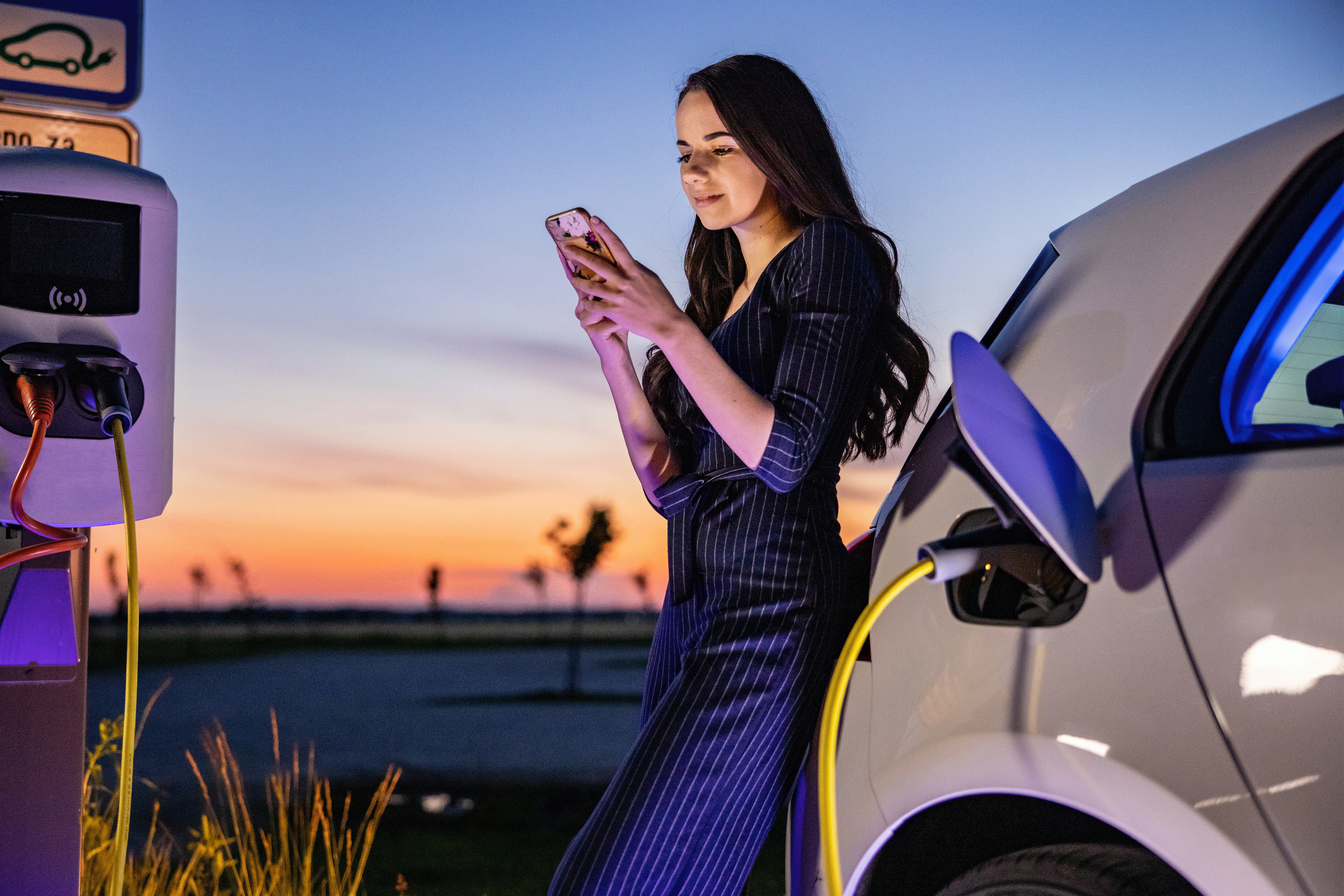 Mujer recarga auto eléctrico mientras mira móvil