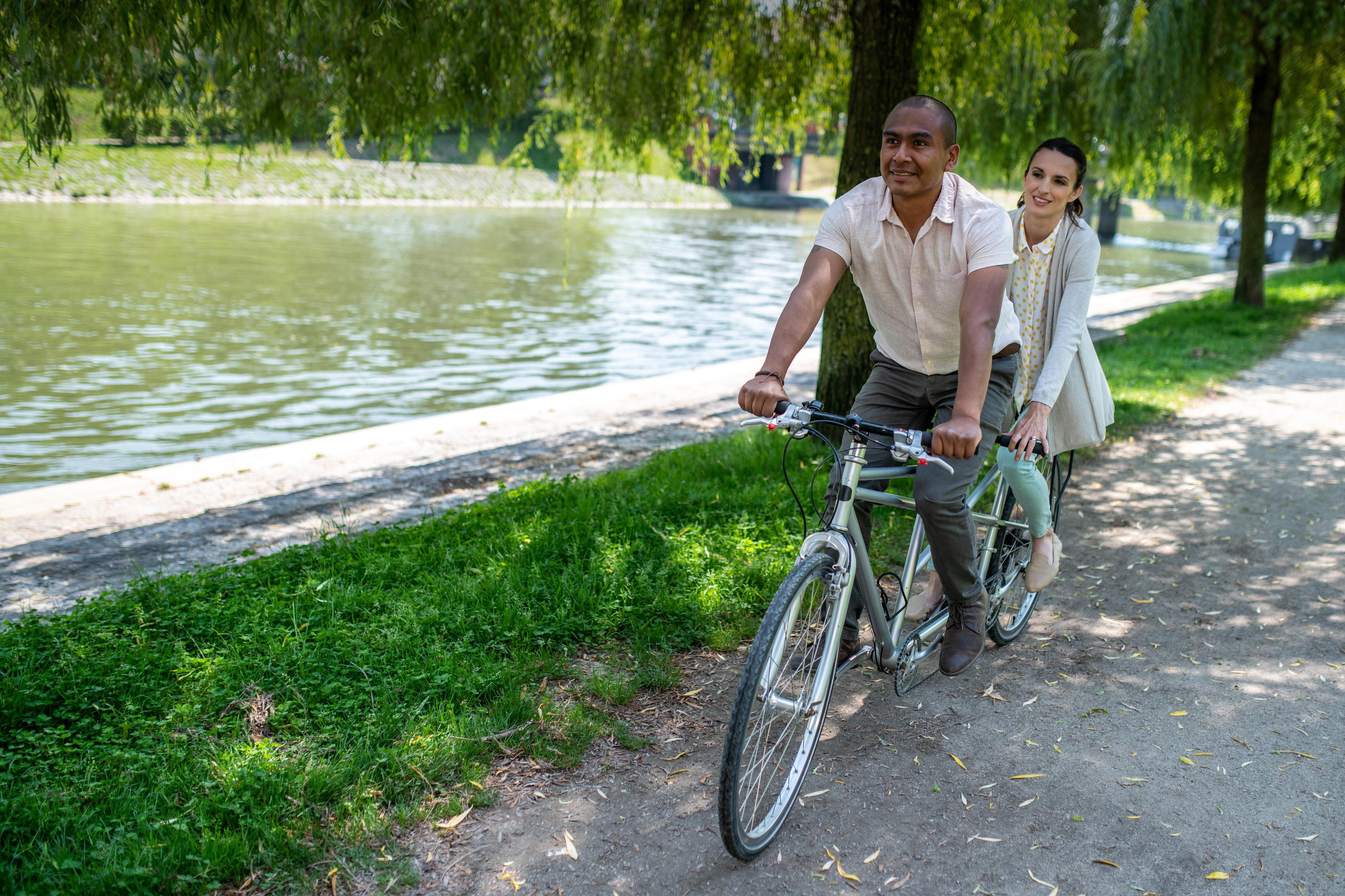 Pareja conduce bici tándem al lado de un río en el parque.