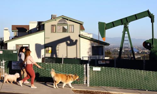 Dos mujeres camina con perros delante de casas y una torre petrolera.